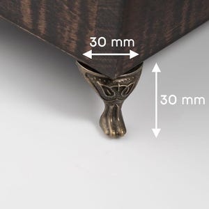 May include: Close-up of a dark brown wooden corner with a decorative metal foot. The foot is 3 cm tall and the corner is 3 cm wide.