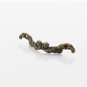 May include: A decorative metal accent piece with a swirling, ornate design. The piece is made of a light gold metal and has a slightly aged finish.