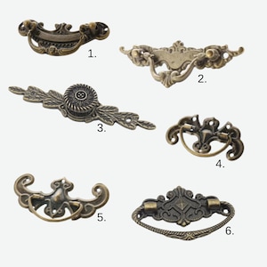 May include: Six antique bronze drawer pulls in various designs. The pulls include ornate scrollwork, floral details, and a variety of handle shapes. Each pull is a different design, offering a selection of hardware options for furniture restoration or new projects.