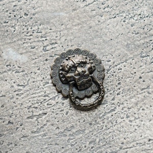 May include: An antique bronze door knocker depicting a lion's head with a ring handle. The knocker is set against a textured, grey surface. The lion's head has detailed features and a weathered look.