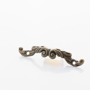 May include: A decorative bronze-colored furniture pull with an ornate, scrolled design. The hardware has two holes for mounting and is curved in shape. Ideal for cabinets, drawers, or other decorative projects.