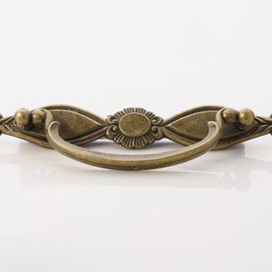 May include: Antique brass-toned drawer pull with a curved handle and ornate detailing. The decorative hardware features a central oval design and stylized floral accents on each end. This vintage-style pull is ideal for cabinets and furniture.
