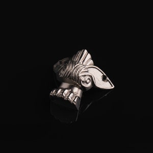 May include: A silver-colored metal pendant in the shape of a stylized head with intricate details. The pendant has a textured surface and a slightly curved shape.