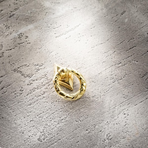 Puede incluir: Un tirador dorado con base en forma de diamante y asa circular. El asa tiene un diseño tejido. La ferretería está colocada sobre una superficie gris texturizada.