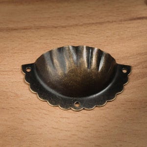 May include: An antique-style drawer pull with a scalloped edge and a bronze finish. The hardware has two holes for mounting and a shell-like design. Suitable for furniture restoration or adding a vintage touch.