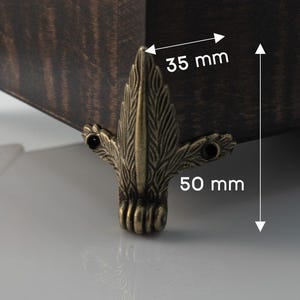 Może przedstawiać: Zbliżenie rogu ciemnobrązowej drewnianej skrzyni z ozdobnym, antycznym, brązowym metalowym narożnikiem. Narożnik ma 3,5 cm szerokości i 5 cm wysokości. Skrzynia ma widoczne usłojenie drewna.