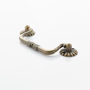 May include: A decorative antique brass cabinet pull with a floral design. The pull has a curved shape and is designed to be used on drawers or doors.