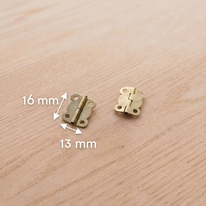 Pode incluir: Duas pequenas dobradiças de cor latão com formato decorativo de borboleta. Cada dobradiça mede aproximadamente 16 mm por 13 mm. As dobradiças são exibidas em uma superfície de madeira clara.