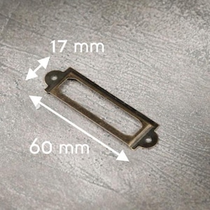 May include: A rectangular, antique brass label holder with a central opening. The label holder is 60 mm long and 17 mm wide. It has rounded edges and two small holes for mounting. The metal has a slightly aged appearance.