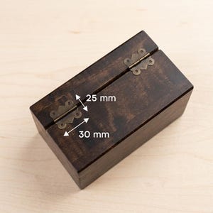 Op de afbeelding: Een donker gebeitste houten kist met een scharnierend deksel en antiek messingkleurige hardware. De kist meet 25 mm bij 30 mm. De kist is rechthoekig en heeft een eenvoudig, klassiek ontwerp.