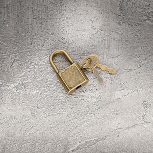 Puede incluir: Un pequeño candado de latón de estilo antiguo con un patrón de diamantes en la parte delantera y dos llaves. El candado y las llaves descansan sobre una superficie gris texturizada.