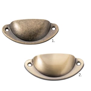 May include: Two antique-style drawer pulls. The top pull has a distressed, bronze finish, while the bottom pull has a polished brass appearance. Both have a cup-shaped design with two mounting holes on each end. These are ideal for furniture restoration.