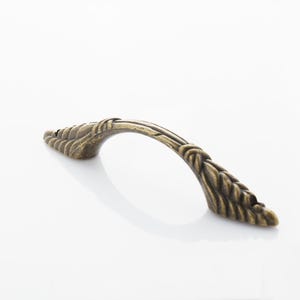 Puede incluir: Un tirador de armario de bronce con un diseño trenzado decorativo. El tirador tiene una forma curva y un extremo puntiagudo.