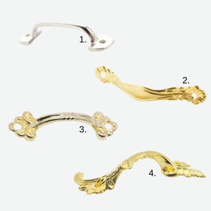 May include: Four cabinet pull handles in different styles and finishes. The handles are numbered 1 to 4. Handle 1 is silver-toned, while handles 2 and 4 are gold-toned with ornate designs. Handle 3 is gold and silver-toned.