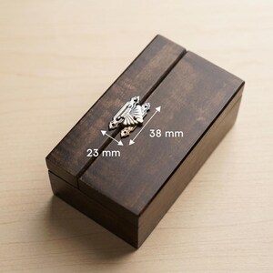May include: A dark brown wooden box with a decorative silver clasp. The box measures 23 mm wide and 38 mm long. The clasp has an ornate design. The box is closed and has a hinged lid.