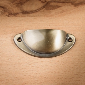 May include: Antique brass-finished cup pull with two mounting holes. The hardware is a half-moon shape and is set against a light brown wooden surface. This cabinet hardware is ideal for drawers and cabinets.