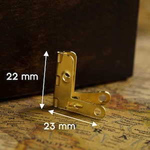 Op de afbeelding: Gouden metalen hoekbeslag met een afmeting van 22 mm bij 23 mm. Het beslag is ontworpen om twee stukken hout of andere materialen in een hoek van 90 graden te verbinden.