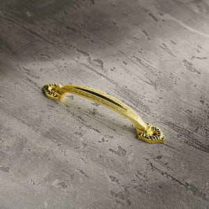 Puede incluir: Un tirador de armario dorado con una superficie texturizada y una franja negra en el centro. El tirador tiene extremos decorativos con detalles ornamentados. El herraje está diseñado para muebles y decoración del hogar.