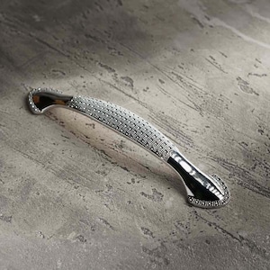 May include: A silver cabinet handle with a curved design. The central section is covered in small, closely spaced, silver-coloured dots. The ends are smooth and curved, with a decorative pattern. The hardware is set against a grey, textured surface.