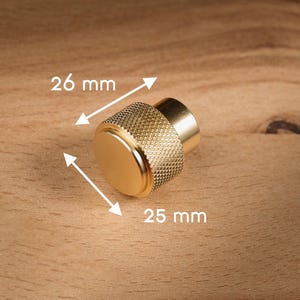 May include: A gold cabinet knob with a textured grip and a flat top. The knob is 2.5 cm in diameter and 2.6 cm long. The hardware is set against a wooden background.