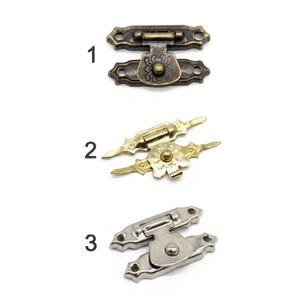 May include: Three metal box latches labeled one, two, and three. The first is an antique bronze finish with floral engraving. The second is a bright gold tone with pointed ends and floral detail. The third is a polished silver tone finish.