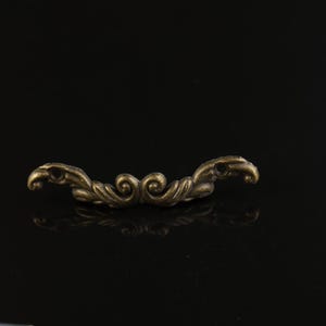 May include: Ornate antique bronze-toned drawer pull with a symmetrical design. The hardware features a curved shape with swirling details and small holes at each end. The pull is set against a black background.