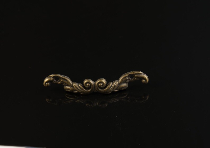 May include: Ornate antique bronze-toned drawer pull with a symmetrical design. The hardware features a curved shape with swirling details and small holes at each end. The pull is set against a black background.