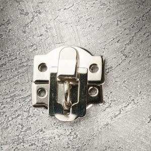 May include: A silver metal latch with a curved top and a hinged closure. The latch has a rectangular base with four screw holes and a central loop for securing. The background is a textured, grey surface.