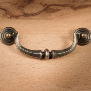 May include: Antique brass drawer pull with a curved handle and circular backplates. The hardware is mounted on a light brown wooden surface. The pull has a vintage aesthetic.