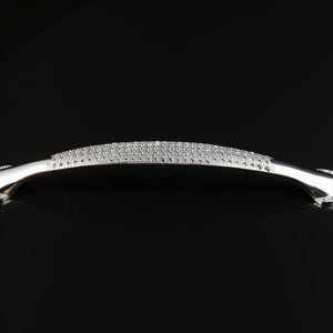 May include: A silver-toned cabinet pull with a curved design. The center section is encrusted with small, sparkling stones, while the ends are outlined with similar stones. The handle is set against a black background.