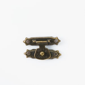 Puede incluir: Un cierre de metal de color bronce con un diseño decorativo. El cierre tiene dos piezas, una con un pomo redondo y la otra con una placa rectangular.