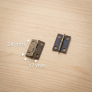 Op de afbeelding: Twee antieke messing scharnieren, elk met vier schroefgaten, worden getoond op een licht houten oppervlak. Eén scharnier is open en de andere is gesloten. Het open scharnier meet 24 mm bij 17 mm.