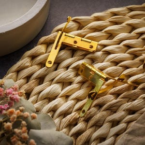 May include: Close-up of two gold-colored metal hinges with a rectangular shape, mounted on a woven, light brown surface. The hinges are positioned at different angles, suggesting they are for a small box or decorative item. A small floral arrangement is in the foreground.