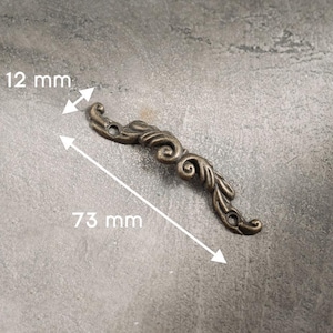 Op de afbeelding: Een decoratief meubelaccent van bronskleurig metaal, 73 mm lang en 12 mm breed. Het sierlijke ontwerp heeft wervelende patronen en twee montagegaten. Het stuk is geplaatst tegen een grijze achtergrond.