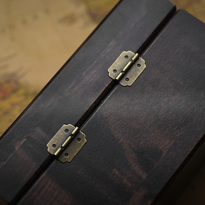 May include: Dark stained wooden box with brass-colored hinges. The box is angled, showing the top and side, with a visible seam and two hinges. The wood grain is visible, and the box appears to be closed.