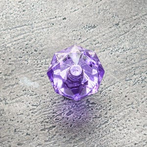 Puede incluir: Una botella de vidrio púrpura con un diseño facetado, que se asemeja a una gema. La botella está vacía y la tapa está quitada.