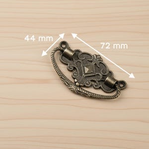 May include: Antique bronze drawer pull with ornate detailing. The pull measures 2.8 inches long and 1.7 inches wide. Features a decorative center with a square accent. Ideal for adding a vintage touch to furniture.