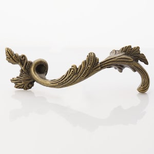May include: An antique-style cabinet handle in a bronze finish. The curved handle is decorated with detailed leaf and scroll patterns. This decorative hardware is likely made of metal and is suitable for furniture restoration or decorative projects.