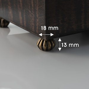 Puede incluir: Primer plano de una caja de madera oscura con patas doradas decorativas. La caja tiene 18 mm de ancho y 13 mm de alto.