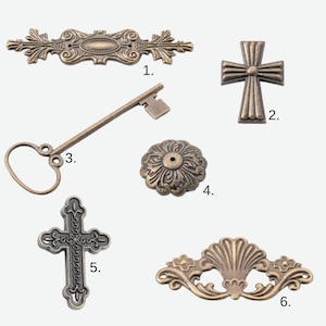 May include: An assortment of antique bronze-colored decorative hardware, including a key, crosses, and ornate drawer pulls. The hardware features intricate floral and geometric designs, suitable for furniture restoration or craft projects.
