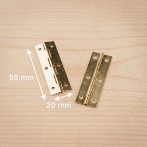 Op de afbeelding: Twee messingkleurige metalen scharnieren, elk met meerdere schroefgaten, worden getoond op een licht houten oppervlak. Eén scharnier is gedeeltelijk open en onthult het vouwmechanisme. De afmetingen zijn 5,8 cm lang en 2 cm breed.