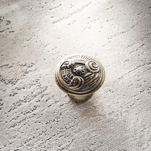 以下が含まれることがあります： 装飾的で渦巻くデザインの銀色のキャビネットノブ。ノブは丸く、詳細なパターンと短いステムが付いています。背景は、テクスチャ加工された明るい灰色の表面です。