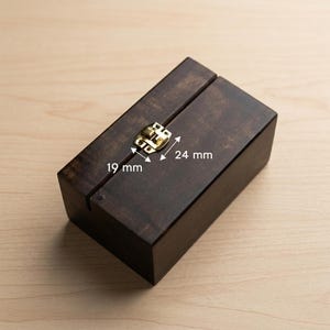 May include: A dark stained wooden box with a brass-colored clasp. The box is rectangular and has a hinged lid. The clasp measures 0.75 inches by 0.94 inches. The box is on a light wood surface.