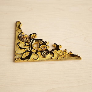 Könnte beinhalten: Ein goldfarbener, verzierter Eckwinkel mit aufwendigen Schnörkeln und einem dunklen, kontrastierenden Hintergrund. Die Halterung hat eine rechtwinklige Form und ist für Möbel oder dekorative Anwendungen konzipiert.