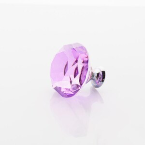 May include: A faceted, purple crystal cabinet knob with a chrome-colored base. The knob is diamond-shaped and reflects light, creating a sparkling effect. The hardware is designed for furniture and home decor.
