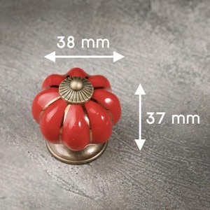 Puede incluir: Pomo de armario de cerámica roja con diseño floral. El pomo mide 37 mm de alto y 38 mm de ancho, con base y detalle central color latón. Imagen de primer plano del herraje decorativo.