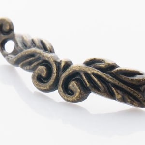 May include: An antique bronze-colored decorative furniture pull with a leaf and scroll design. The hardware has two holes for mounting and is slightly curved. The metal has a distressed finish.