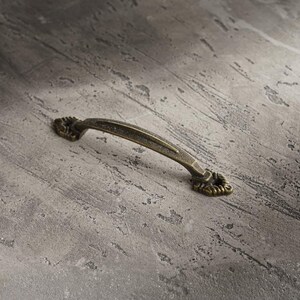May include: An antique-style bronze drawer pull with decorative floral accents. The curved handle is set against a textured gray surface, creating a vintage aesthetic. Ideal for cabinets and furniture.