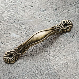 May include: An antique-style bronze cabinet pull with ornate detailing. The handle features a curved design with decorative flourishes at each end, and a central grip area. The hardware is a warm, aged bronze color, suggesting a vintage aesthetic.