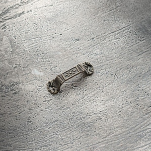 Puede incluir: Un pequeño tirador de cajón de metal adornado con un diseño decorativo. El tirador es de color bronce claro y tiene un acabado ligeramente envejecido.